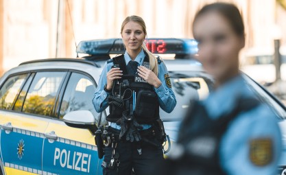 Zwei Polizistinnen stehen vor einem Streifenwagen. Dabei liegt der Fokus auf der Beamtin in blauer Polizeiuniform in der Mitte. Sie hat blonde, lange Haare und schaut in die Kamera.