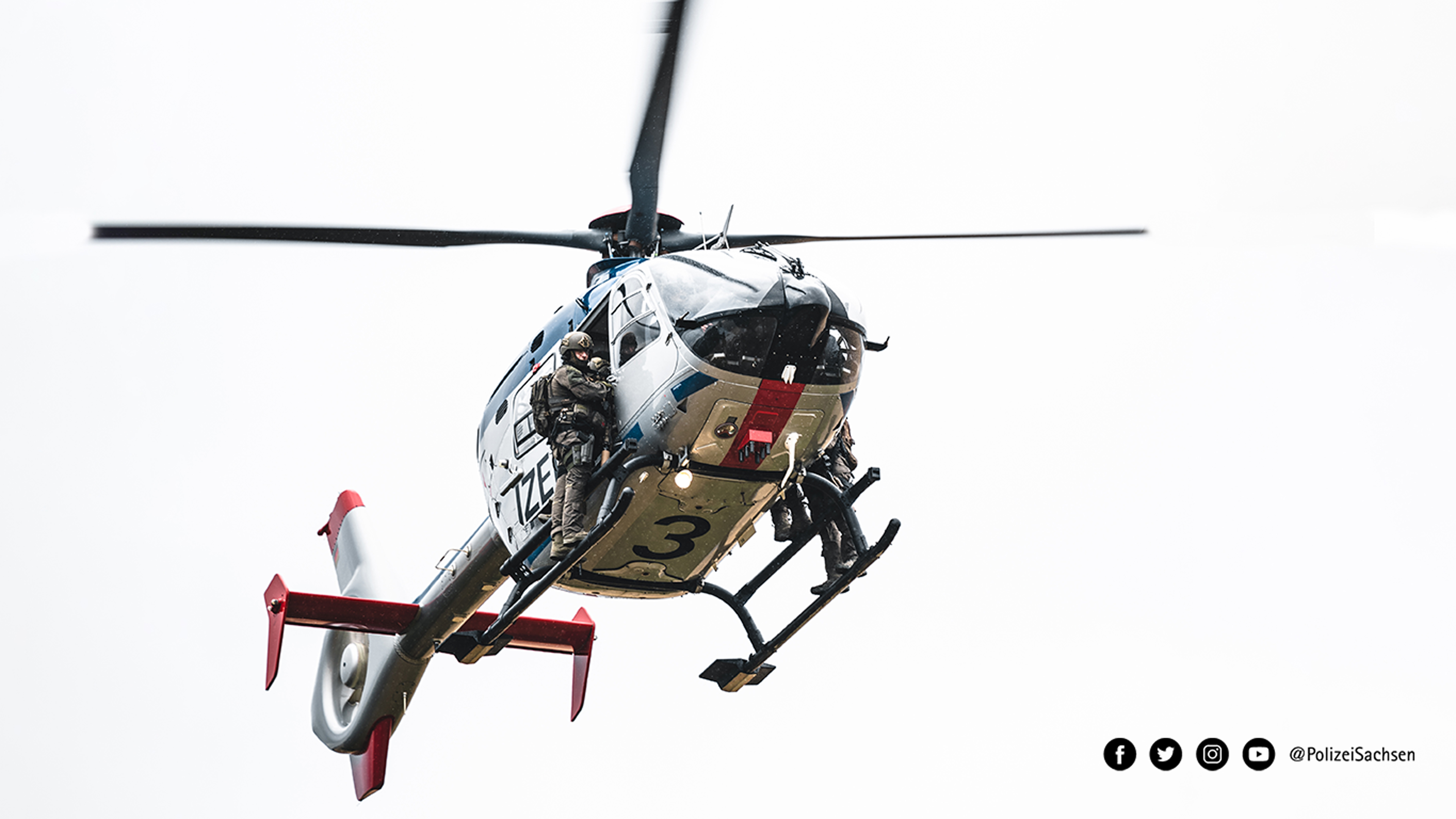 Ein Hubschrauber der Polizei Sachsen in der Luft. SEK-Beamte stehen auf den Kufen und halt sich fest.