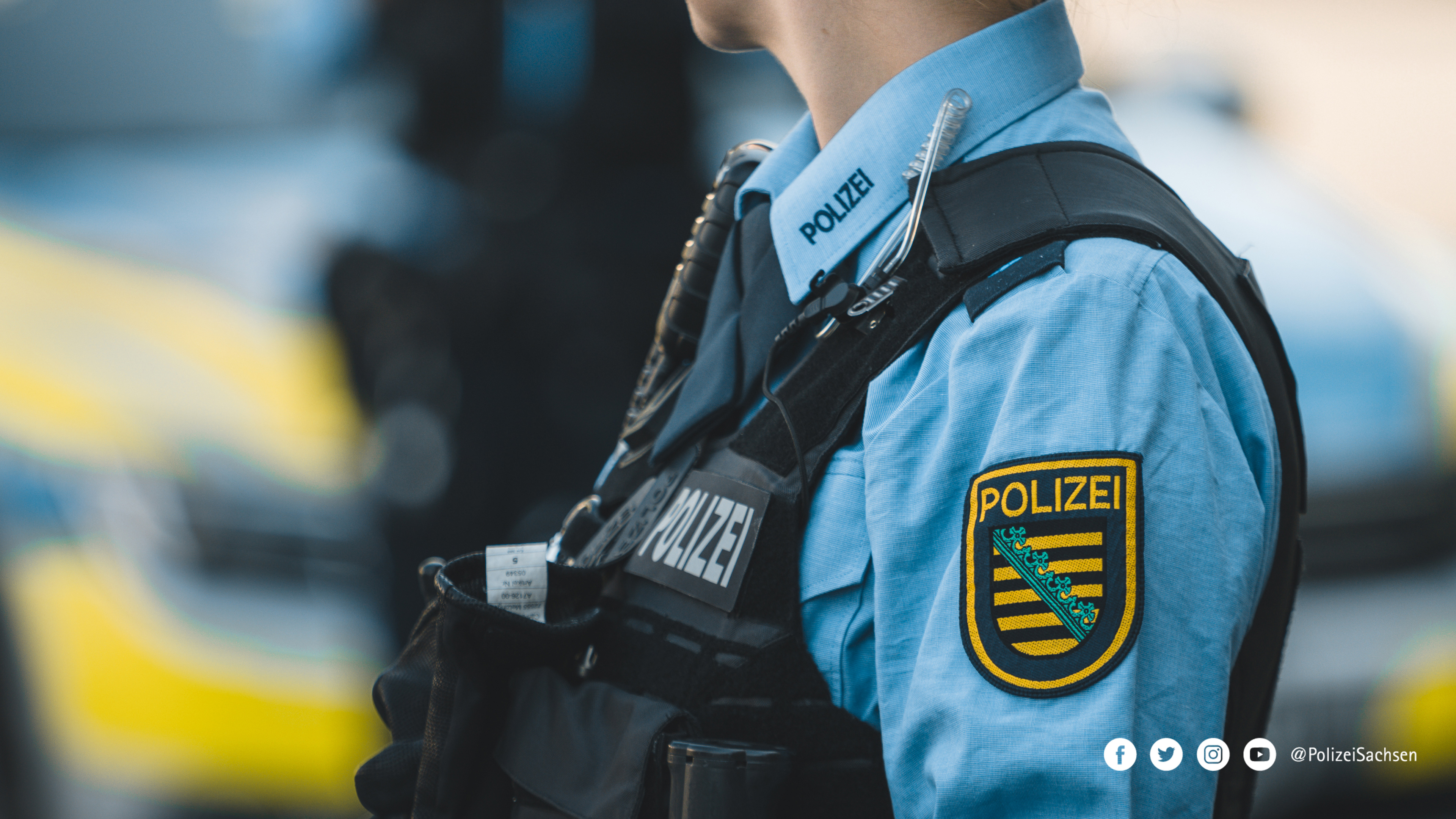 Eine Streifenbeamtin von der Seite: Zu sehen sind die Schutzweste, das Funkgerät, das Hemd und der Aufnäher der Polizei Sachsen.