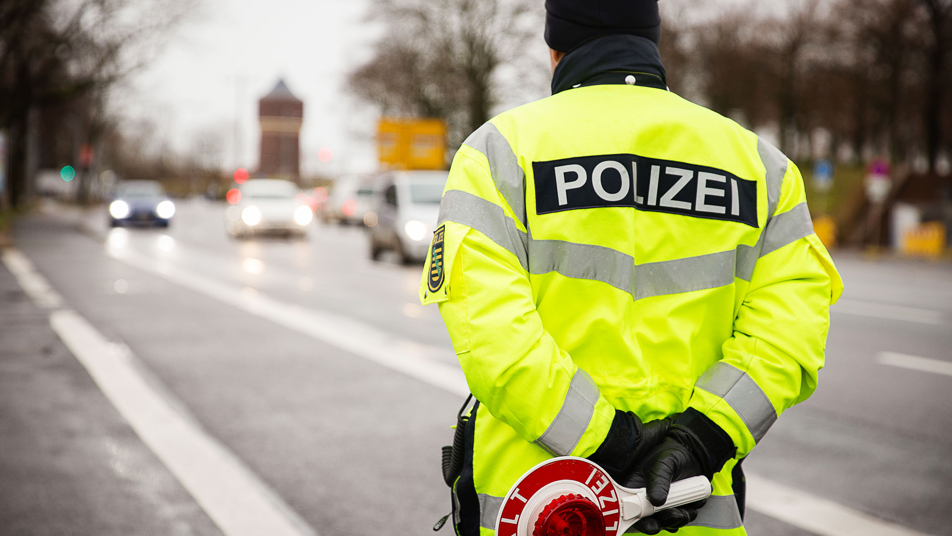Ein Polizist mit neongelber Jacke steht am Straßenrand und beobachtet den Verkehr. Er hält hinter seinem Rücken eine Kelle.